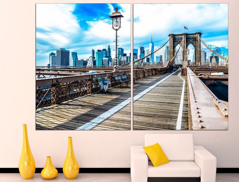 May include: A split canvas print featuring a view of the Brooklyn Bridge in New York City. The bridge is a suspension bridge with a brown wooden walkway and a view of the city skyline in the background. The sky is blue with white clouds.