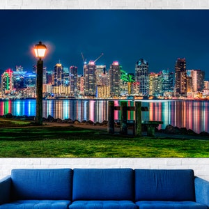 Puede incluir: Una vista nocturna de un horizonte urbano con una sola farola y un banco de madera en primer plano. La ciudad est&aacute; iluminada con luces de colores que se reflejan en el agua.