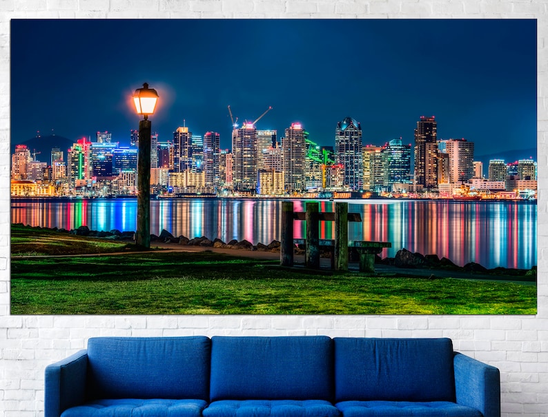 Puede incluir: Una vista nocturna de un horizonte urbano con una sola farola y un banco de madera en primer plano. La ciudad est&aacute; iluminada con luces de colores que se reflejan en el agua.