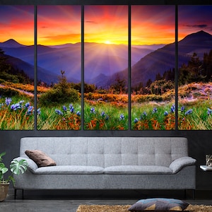 May include: A panoramic landscape photograph of a mountain range at sunset. The sky is a vibrant orange and pink, with the sun setting behind the mountains. The mountains are covered in lush green vegetation and there are purple wildflowers in the foreground.