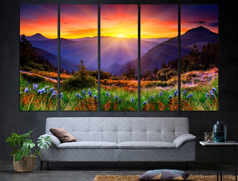 May include: A panoramic landscape photograph of a mountain range at sunset. The sky is a vibrant orange and pink, with the sun setting behind the mountains. The mountains are covered in lush green vegetation and there are purple wildflowers in the foreground.