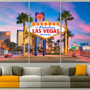 May include: A triptych canvas print featuring the iconic "Welcome to Fabulous Las Vegas Nevada" sign at dusk. The sign is illuminated against a backdrop of palm trees, buildings, and a vibrant sky. The artwork is displayed above a modern sofa.