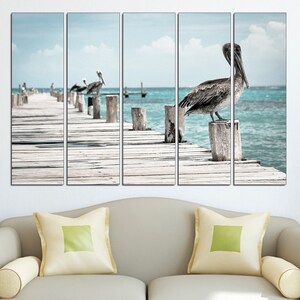 May include: A multi-panel canvas print featuring a pelican perched on a wooden post over a turquoise ocean. The pelican is brown and gray with a long beak. The wooden post is weathered and gray. The ocean is calm and blue. The background is a blue sky with white clouds.