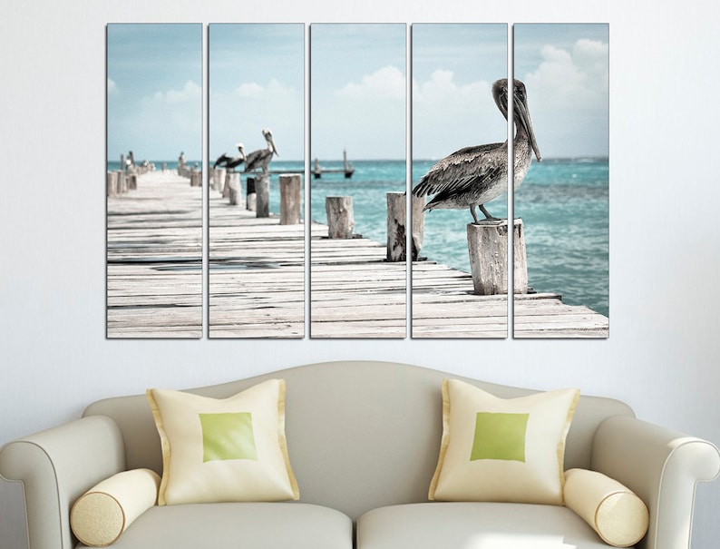 May include: A multi-panel canvas print featuring a pelican perched on a wooden post over a turquoise ocean. The pelican is brown and gray with a long beak. The wooden post is weathered and gray. The ocean is calm and blue. The background is a blue sky with white clouds.