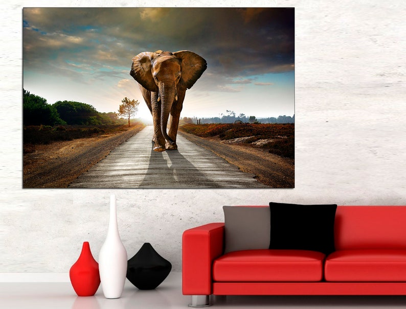 May include: A large brown elephant walks down a dirt road towards the sunset. The elephant is in the middle of the road, with a long shadow behind it. The road is surrounded by green trees and bushes.