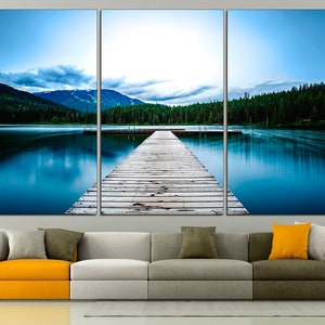 May include: A wooden dock extends out into a still blue lake, surrounded by a lush green forest and snow-capped mountains in the distance. The sky is a light blue with white clouds.