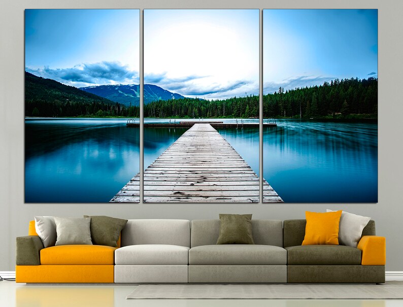 May include: A wooden dock extends out into a still blue lake, surrounded by a lush green forest and snow-capped mountains in the distance. The sky is a light blue with white clouds.