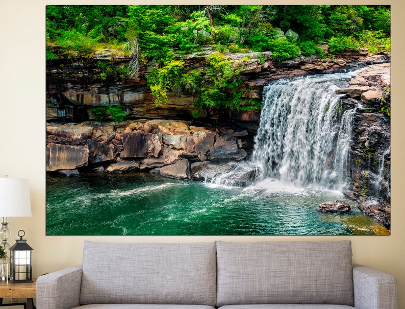 May include: A cascading waterfall flows over moss-covered rocks into a clear, green pool. Lush greenery surrounds the waterfall, creating a serene and picturesque scene.