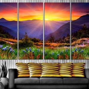 May include: A multi-panel canvas print depicting a vibrant sunset over a mountain range. The sky is a mix of orange, pink, and purple, with rays of light emanating from the sun. The mountains are a deep blue, and the foreground is a lush green meadow with wildflowers.