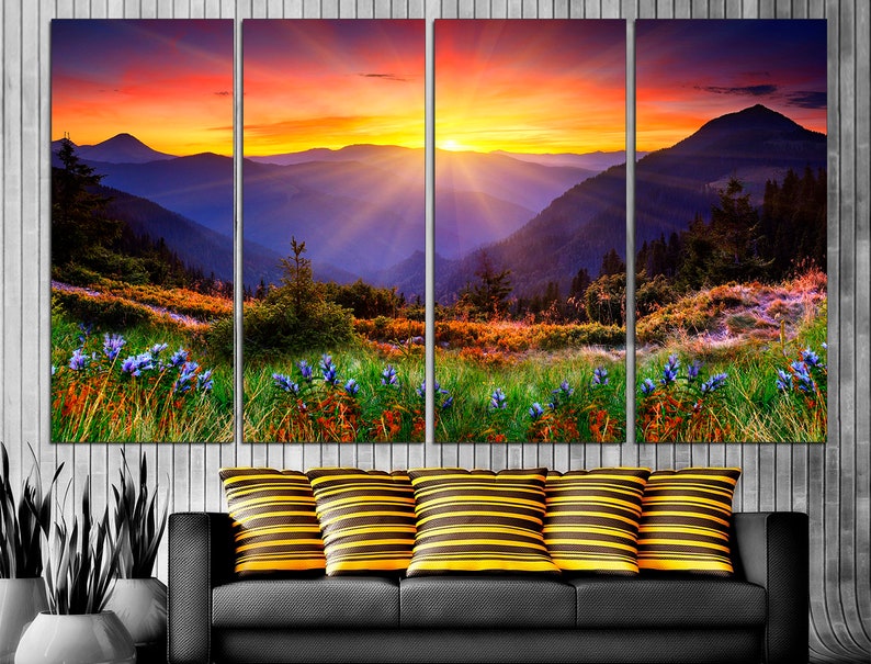 May include: A multi-panel canvas print depicting a vibrant sunset over a mountain range. The sky is a mix of orange, pink, and purple, with rays of light emanating from the sun. The mountains are a deep blue, and the foreground is a lush green meadow with wildflowers.