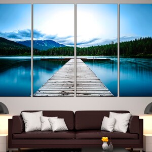 May include: A wooden dock extends out into a still blue lake, surrounded by a lush green forest and mountains in the distance. The sky is a soft blue with wispy clouds.