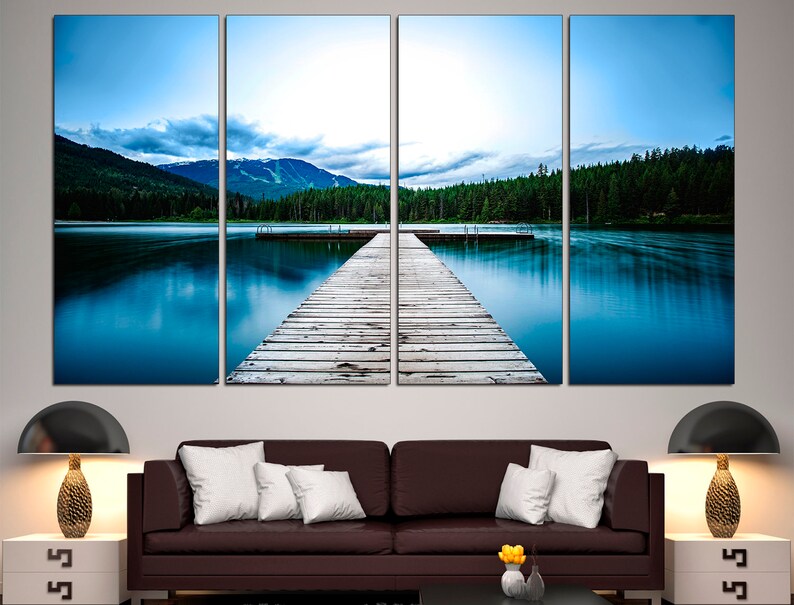 May include: A wooden dock extends out into a still blue lake, surrounded by a lush green forest and mountains in the distance. The sky is a soft blue with wispy clouds.
