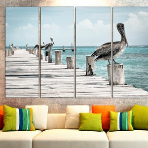 May include: A multi-panel canvas print featuring a pelican perched on a wooden post over a turquoise ocean. The pelican is facing the right side of the image. The wooden post is part of a wooden pier extending into the ocean. The background is a blue sky with white clouds.