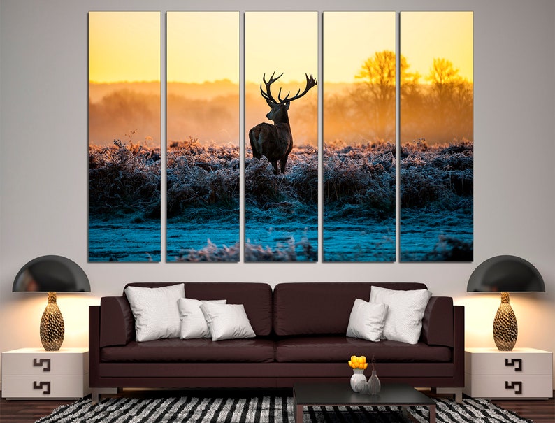 May include: A multi-panel canvas print depicting a silhouette of a deer standing in a field at sunrise. The deer is facing away from the viewer, with its antlers visible. The background is a soft, golden light, with a hint of blue in the distance.