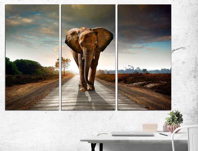 May include: A large brown elephant walks down a dirt road in a field. The sky is cloudy and the sun is setting in the distance.