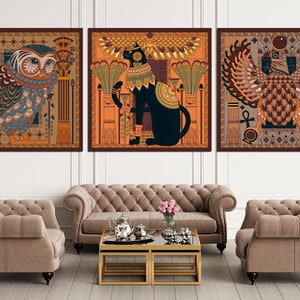 May include: Three square framed prints with an Egyptian theme. The first print features a blue and brown owl with intricate details. The second print features a black cat with a golden collar and a red background. The third print features a brown and gold winged creature with a red background.