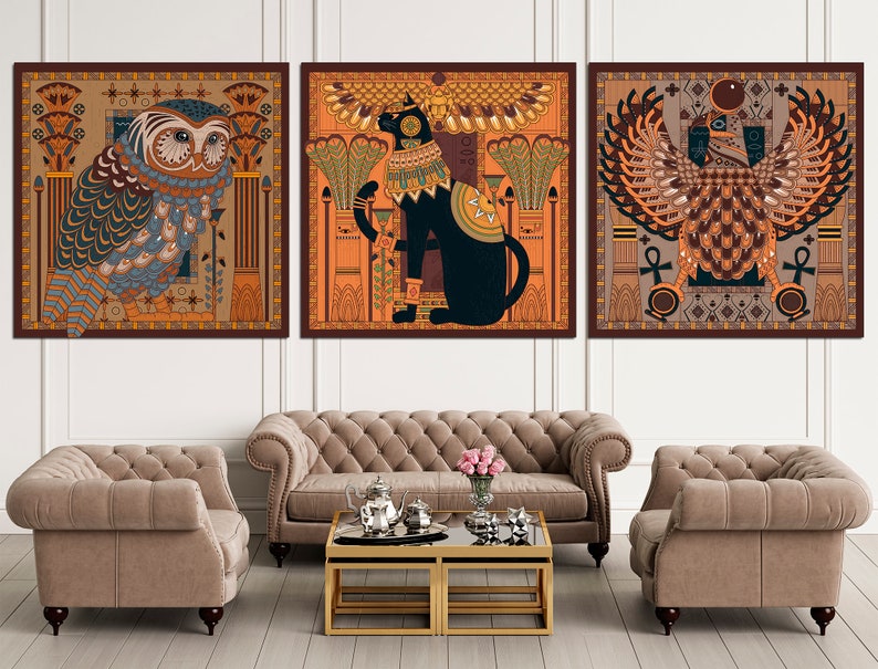 May include: Three square framed prints with an Egyptian theme. The first print features a blue and brown owl with intricate details. The second print features a black cat with a golden collar and a red background. The third print features a brown and gold winged creature with a red background.