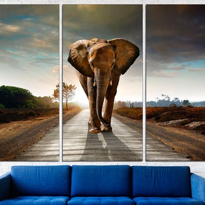 May include: A large brown elephant walks down a dirt road towards the sunset. The elephant is in the middle of the road, with trees and bushes on either side. The sky is a mix of blue and gray clouds.