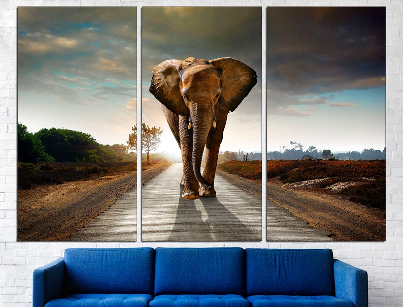 May include: A large brown elephant walks down a dirt road towards the sunset. The elephant is in the middle of the road, with trees and bushes on either side. The sky is a mix of blue and gray clouds.