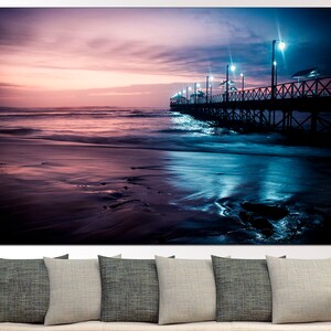 May include: A canvas print of a beach scene at dusk. The image features a wooden pier extending into the ocean, illuminated by lights. The sky transitions from pink to blue, reflecting in the wet sand and water.