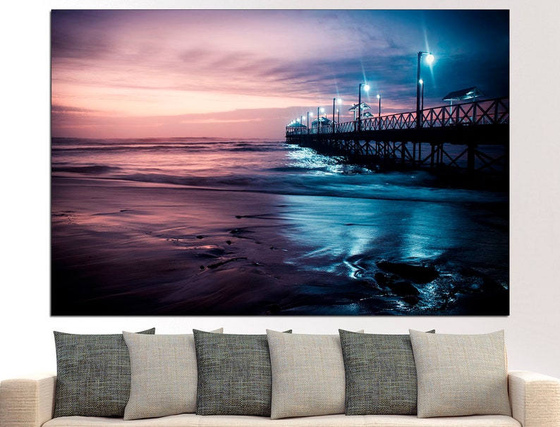 May include: A canvas print of a beach scene at dusk. The image features a wooden pier extending into the ocean, illuminated by lights. The sky transitions from pink to blue, reflecting in the wet sand and water.