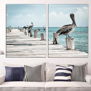 May include: A split canvas print featuring a wooden dock extending over turquoise water. A pelican stands on a post at the end of the dock, while other pelicans are perched on posts further down the dock.