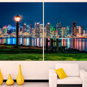 Puede incluir: Una ciudad nocturna con un horizonte urbano reflejado en el agua. Una sola farola ilumina el primer plano, que presenta una zona de c&eacute;sped y un banco de madera.