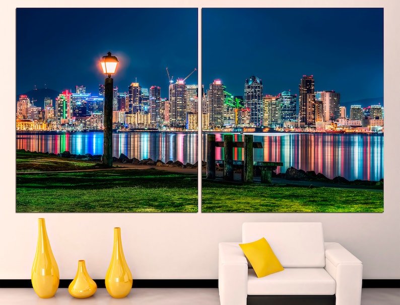 Puede incluir: Una ciudad nocturna con un horizonte urbano reflejado en el agua. Una sola farola ilumina el primer plano, que presenta una zona de c&eacute;sped y un banco de madera.