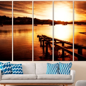 May include: A sunset over a body of water with a wooden pier extending into the water. The sky is a vibrant orange and yellow, and the water is a deep blue. There are boats in the distance.