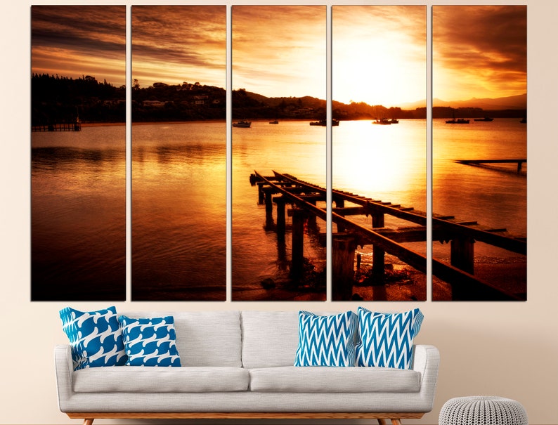 May include: A sunset over a body of water with a wooden pier extending into the water. The sky is a vibrant orange and yellow, and the water is a deep blue. There are boats in the distance.