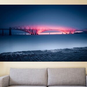 May include: A red-lit bridge over a body of water with a cobblestone foreground. The bridge is a steel truss bridge with a dark blue sky and fog in the background.