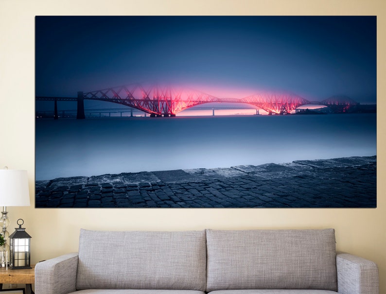 May include: A red-lit bridge over a body of water with a cobblestone foreground. The bridge is a steel truss bridge with a dark blue sky and fog in the background.