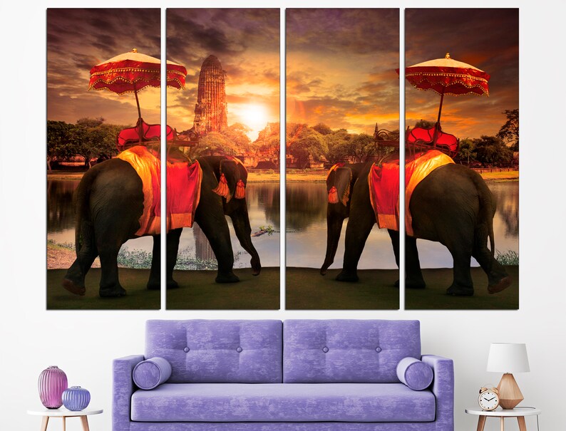 May include: Two elephants adorned with colorful fabric and parasols walk along a riverbank at sunset. The elephants are in front of a temple with a golden spire. The scene is a vibrant depiction of Thai culture.