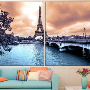 May include: A split canvas print featuring a panoramic view of the Eiffel Tower in Paris, France. The image shows the tower in the distance, with a bridge and river in the foreground. The sky is a soft orange and pink, and the water is a cool blue.