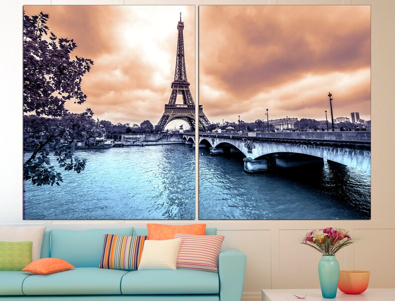 May include: A split canvas print featuring a panoramic view of the Eiffel Tower in Paris, France. The image shows the tower in the distance, with a bridge and river in the foreground. The sky is a soft orange and pink, and the water is a cool blue.