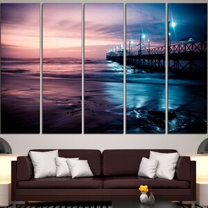 May include: A multi-panel canvas print depicting a serene beach scene at dusk. The artwork features a pier extending into the ocean, illuminated by streetlights. The sky transitions from pink to deep blue, reflecting on the water and wet sand.