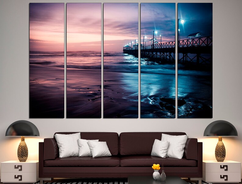 May include: A multi-panel canvas print depicting a serene beach scene at dusk. The artwork features a pier extending into the ocean, illuminated by streetlights. The sky transitions from pink to deep blue, reflecting on the water and wet sand.