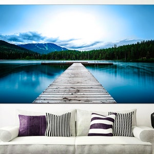 May include: A wooden dock extends out into a still blue lake, with a mountain range in the background and a forest on the opposite shore.