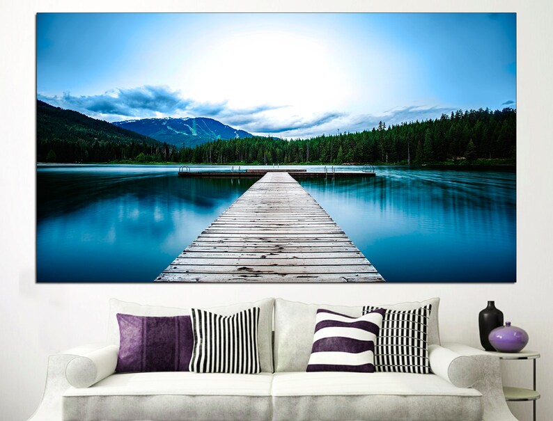 May include: A wooden dock extends out into a still blue lake, with a mountain range in the background and a forest on the opposite shore.