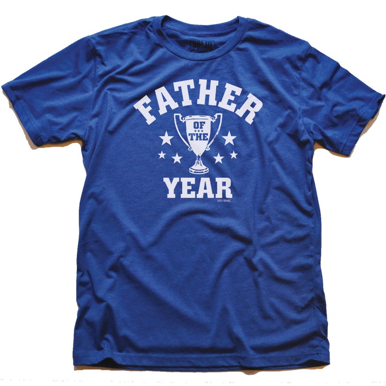 May include: A royal blue short-sleeved t-shirt featuring a white graphic print on the chest. The design includes the text FATHER OF THE YEAR in a bold font, centered around an illustration of a trophy cup surrounded by six small stars.