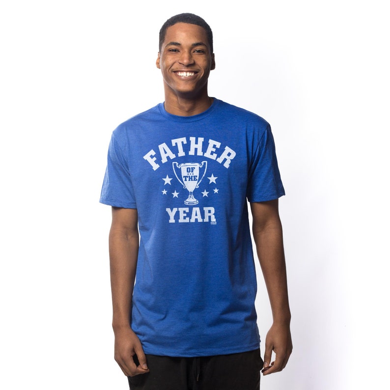 May include: A royal blue short-sleeved t-shirt featuring a white graphic print on the chest. The design includes the text FATHER OF THE YEAR in a bold, arched font, centered above and below a trophy icon surrounded by six small white stars.