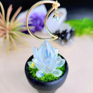 Puede incluir: Una escultura decorativa de cristal con una luna creciente dorada y un pequeño colgante de cristal iridiscente. La escultura presenta un gran cristal facetado en una maceta negra con musgo verde. El cristal refleja la luz.