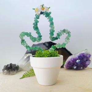 Pode incluir: Uma escultura de cacto artesanal feita de lascas de pedras preciosas verdes, adornada com uma coroa floral, está em um pequeno vaso branco cheio de musgo verde. A escultura é exibida em uma superfície de madeira, um item decorativo único.