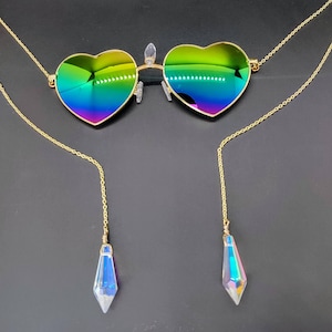 May include: Heart-shaped sunglasses with a gold frame and iridescent lenses. The glasses are connected to a gold chain and feature dangling crystal pendants. The lenses reflect a rainbow of colors, creating a vibrant and eye-catching accessory.