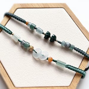 May include: Two jade bracelets with green and blue beads and a dark green woven cord. The bracelets are displayed on a wooden hexagonal tray with a white interior. The beads are various shapes and sizes.