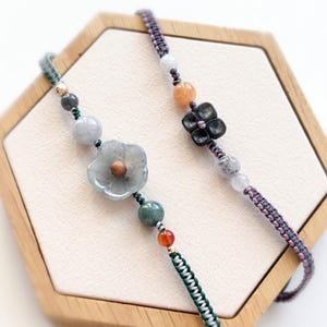 Op de afbeelding: Twee handgemaakte armbanden met bloemen- en kralenontwerpen. Eén armband heeft een grijze bloem en groene kralen, terwijl de andere een zwarte bloem en oranje kralen heeft. De armbanden worden gepresenteerd op een houten zeshoekige doos.