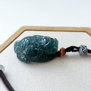 May include: A dark teal jade turtle pendant with intricate carved details. The turtle is attached to a black cord with a wooden bead and a decorative knot. The pendant is displayed on a light-colored surface.