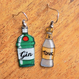 Op de afbeelding: Een paar oorbellen in de vorm van flessen, een groene met het woord "Gin" en de andere gele met het woord "Tonic".