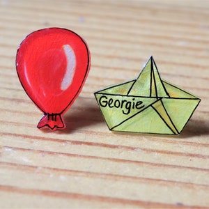 May include: Two acrylic pins: a red balloon with a black outline and a yellow paper boat with the name "Georgie" written on it. The pins are on a wooden surface.