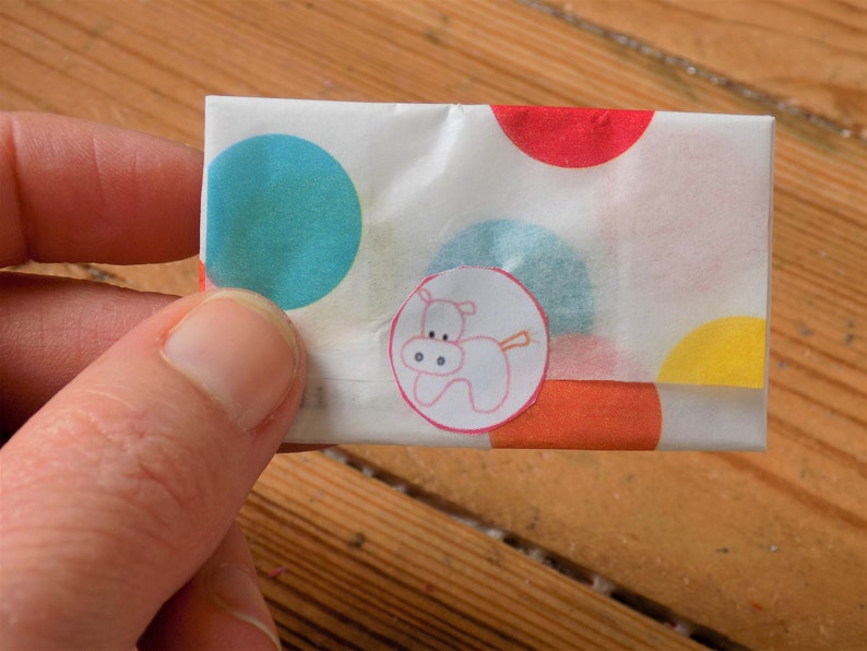 Puede incluir: Un sobre de papel blanco con lunares de colores y una pegatina de un hipop&oacute;tamo rosa. El sobre est&aacute; doblado por la mitad y tiene un patr&oacute;n de lunares rojos, naranjas, azules y amarillos.